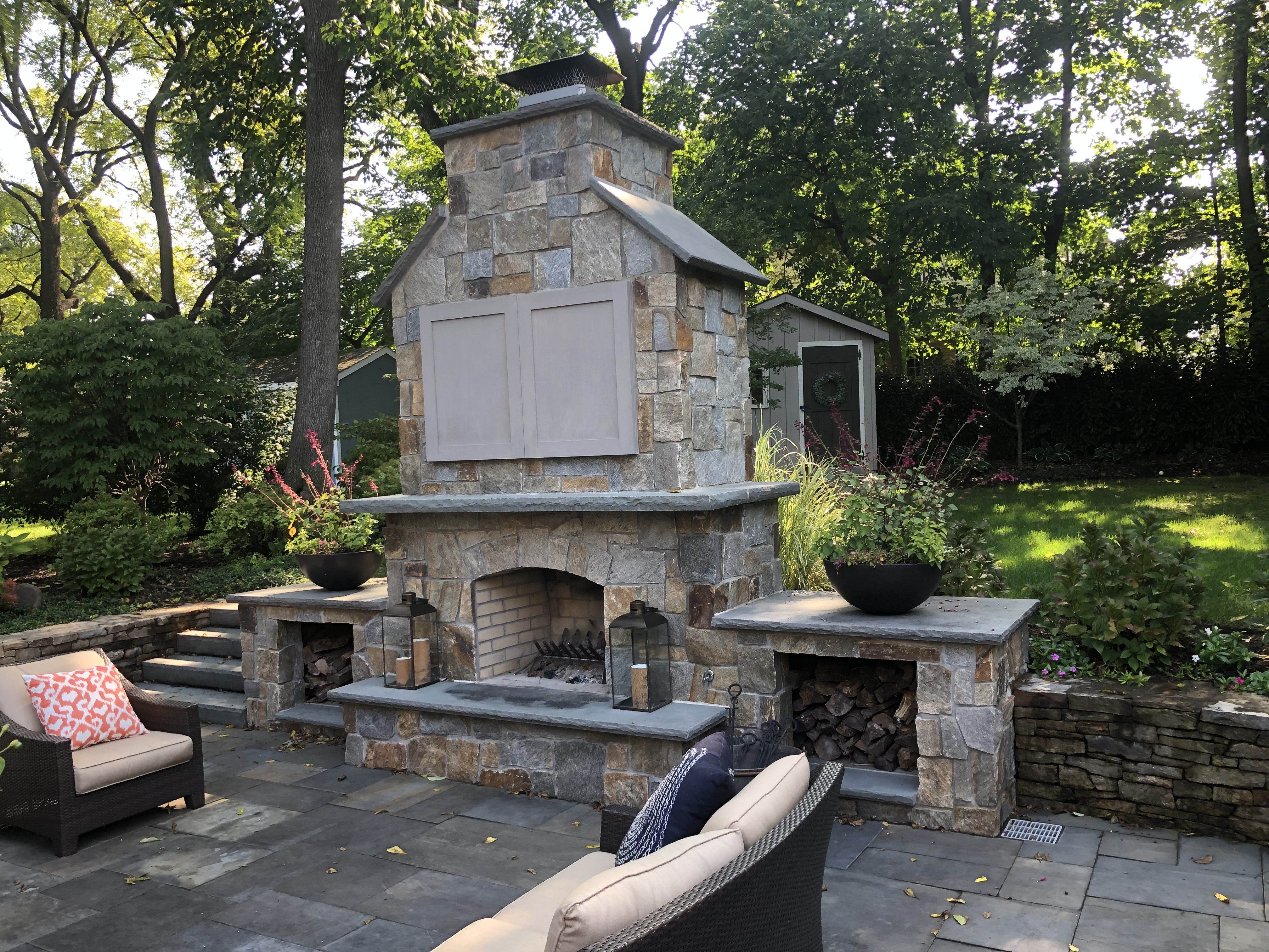 Outdoor stone fireplace with firewood storage, lanterns and lounge seating on bluestone patio