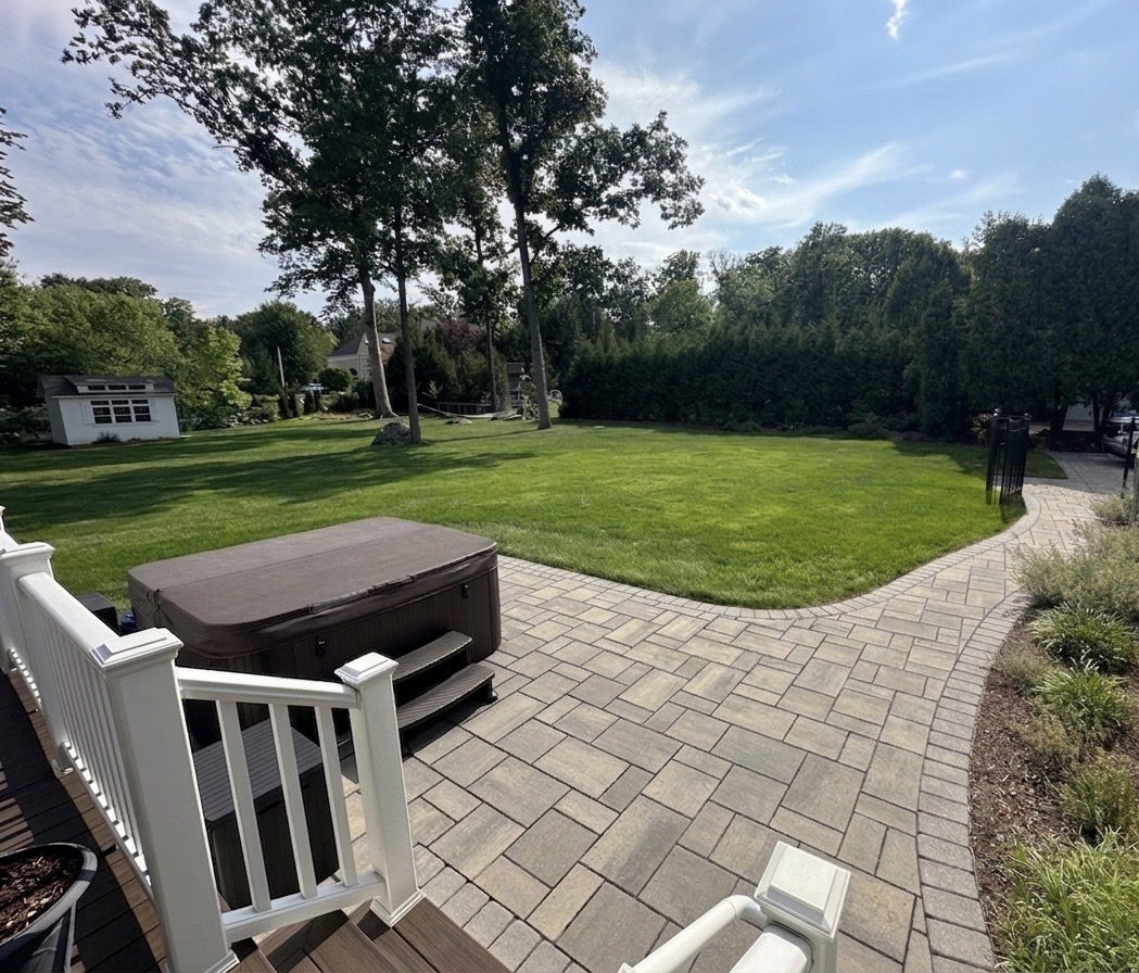 Curved paver patio with hot tub pad, white deck railing and expansive manicured rear lawn