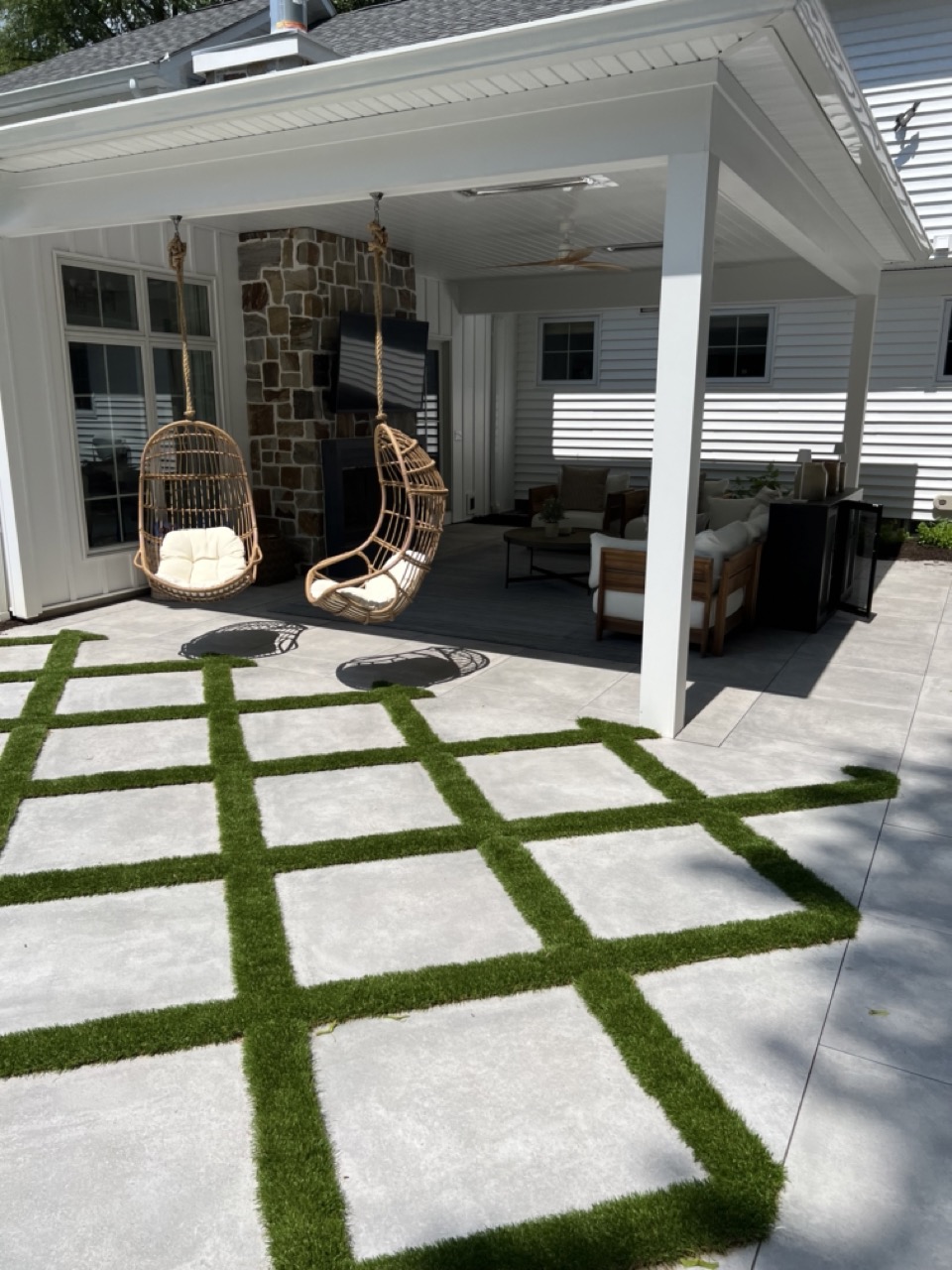 Covered outdoor living room with stone fireplace, hanging rattan chairs and large-format paver/turf grid — Wyckoff